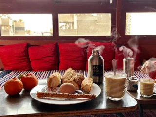 A breakfast table with bread, boiled egg, cinnamon stick, tomatoes, and steaming drinks on a tray. Red cushions are in the background.