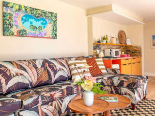 A living room with a floral-patterned sofa, wooden coffee table, and kitchenette in the background. A colorful painting hangs on the wall.