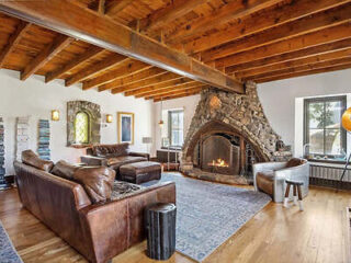 A cozy living room with wooden beams, stone fireplace, and leather sofas. A large rug covers the wooden floor.