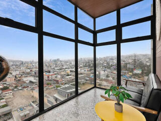 A room with large glass windows offers a panoramic city view. A black couch and a yellow table with a plant are in the foreground.