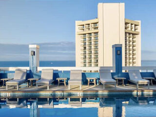 Rooftop pool with lounge chairs and a view of a tall building and ocean in the background.