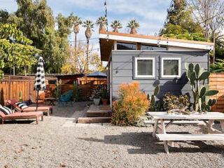 A modern small gray house with a gravel yard, white picnic table, sun loungers, potted plants, and string lights, surrounded by a wooden fence and tall trees.