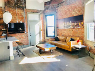 A loft-style living room with exposed brick walls, a brown leather couch, a coffee table, and a wall-mounted TV. Large windows let in natural light.