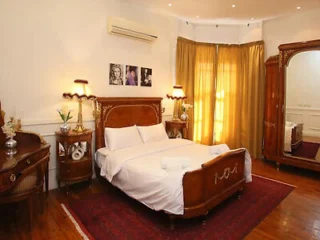 A bedroom with a wooden bed, white bedding, matching side tables, a vanity desk, and a large mirror. A red rug covers the wooden floor. Bright curtains and framed photos are on the walls.