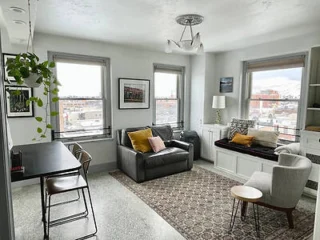 A bright living room with a gray sofa, a window seat, and a small dining table. Large windows reveal a cityscape, while plants and framed pictures decorate the space.