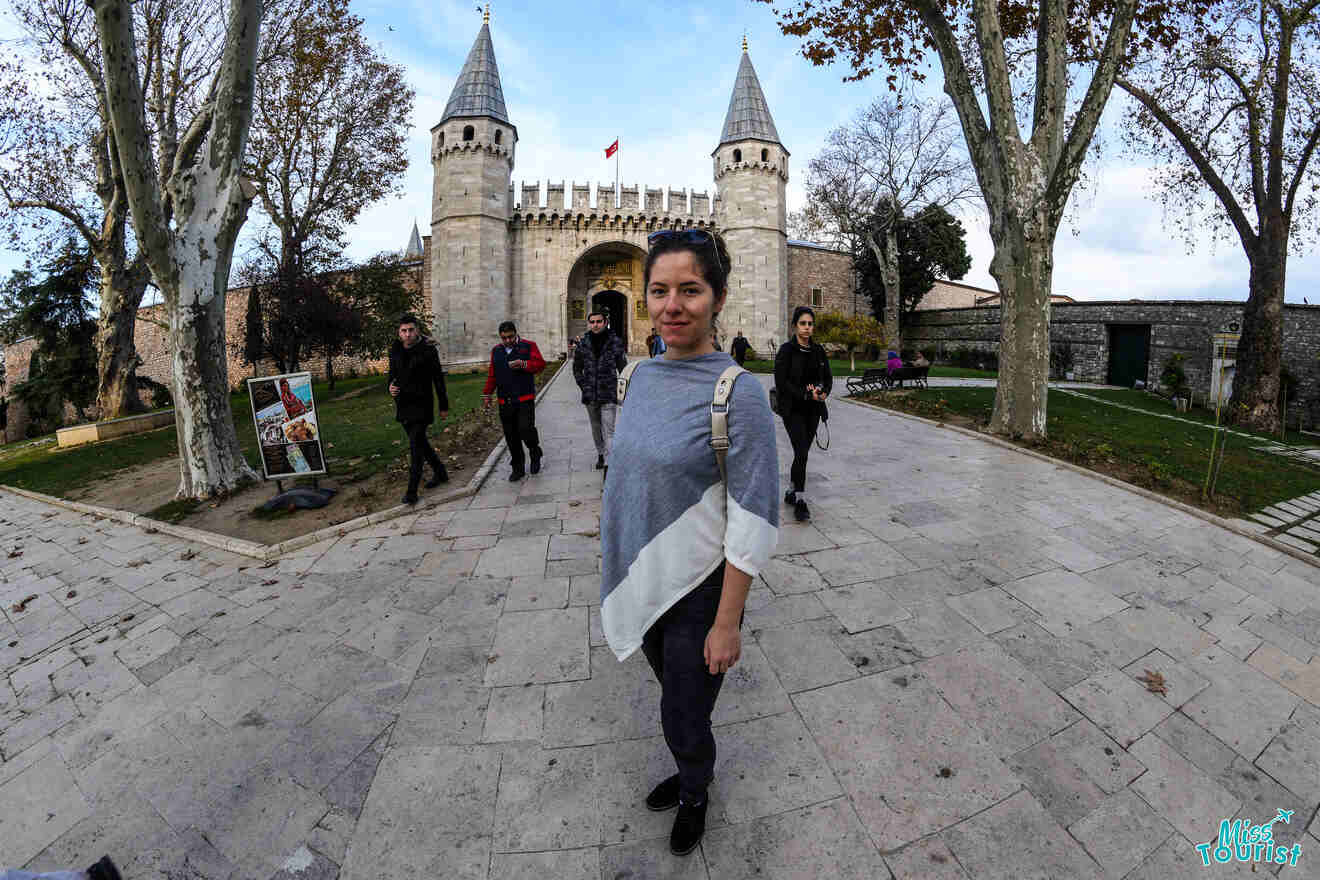 author of the post stands in front of a historical gate with two towers. Other people walk around, and trees are in the background.