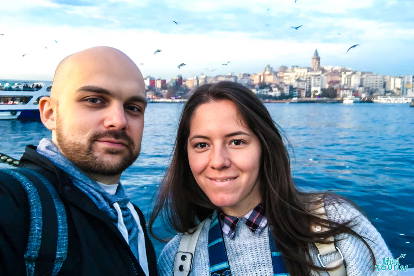 The writer of the post with her husband take a selfie by the water with a cityscape and seagulls in the background.