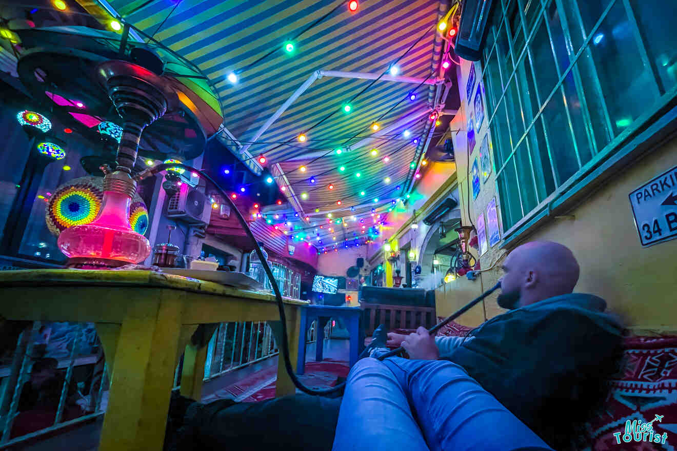 A person relaxes on a cushioned seat under vibrant, multicolored string lights in a cozy, tent-like space with a visible hookah on the table.