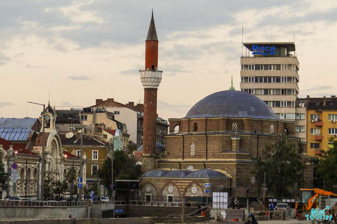 A cityscape featuring a mosque with a tall minaret and a dome, surrounded by various buildings and trees under a cloudy sky.