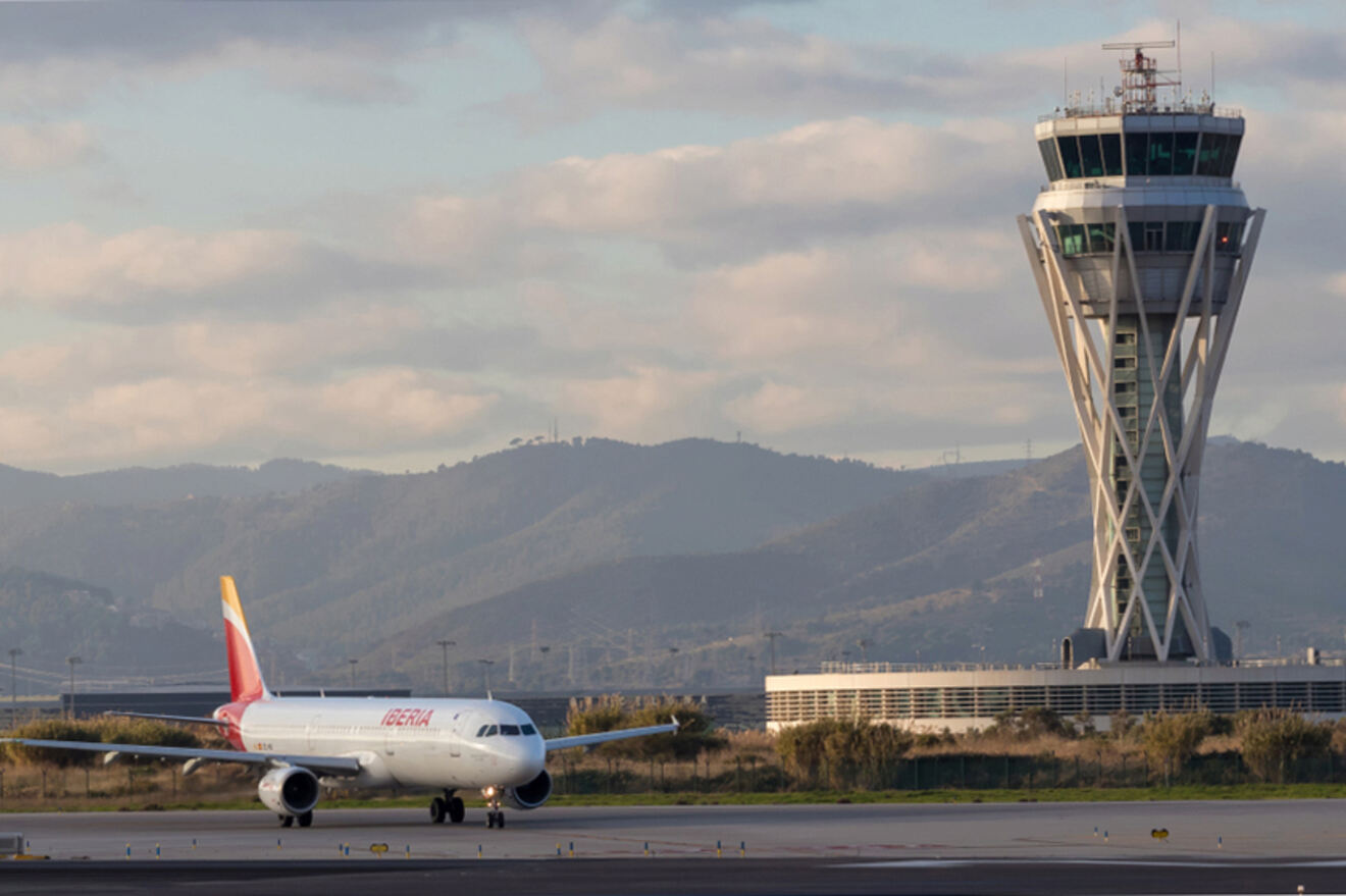 A commercial airplane on the runway near an airport control tower, with mountains in the background.