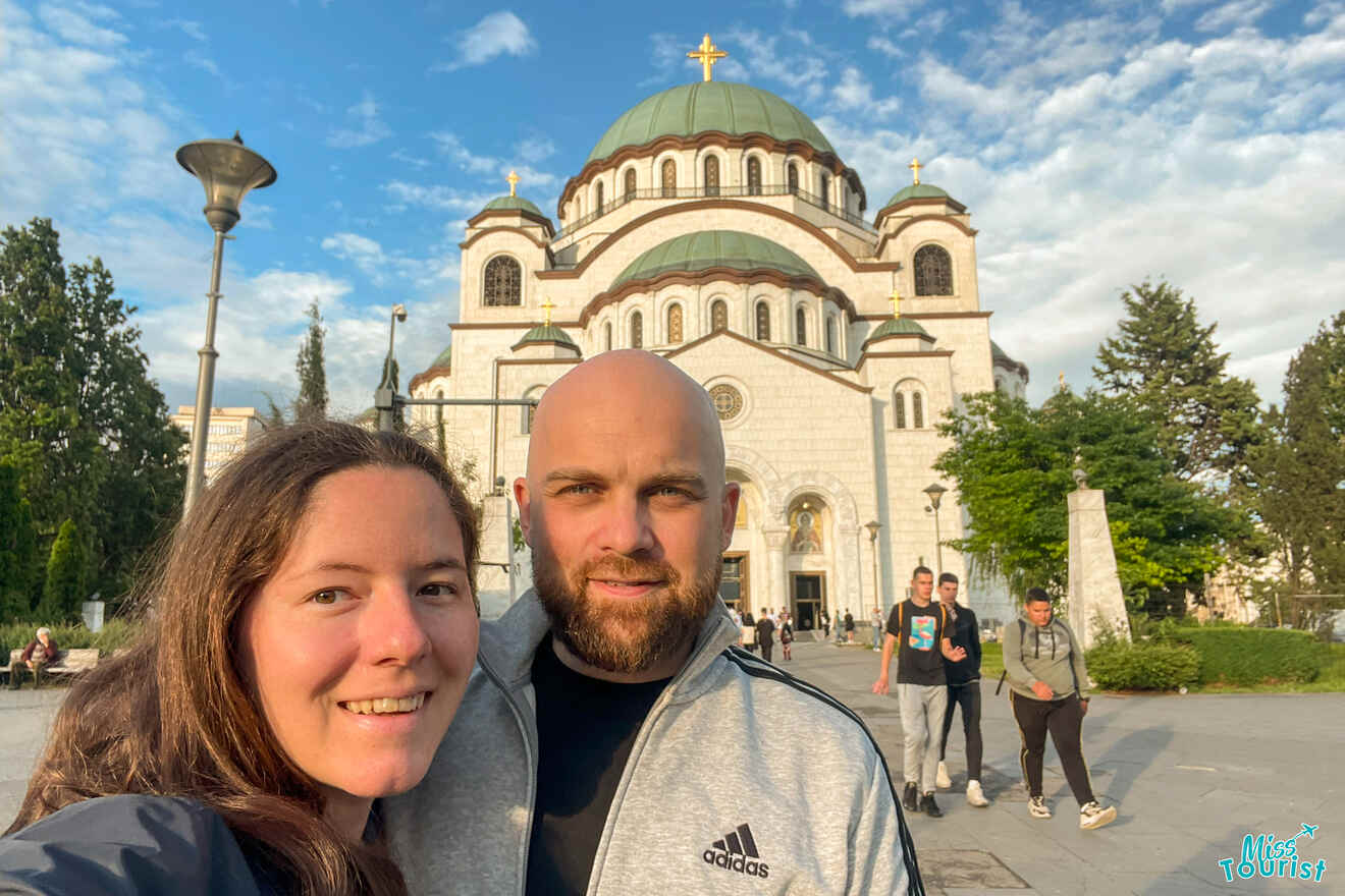 On their Serbia itinerary, writer of the post with her husband takes a selfie in front of a majestic white Orthodox church with a green dome and gold cross, surrounded by trees and strolling visitors.