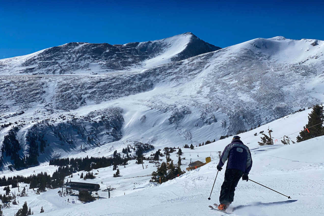 A skier descends a snowy mountain slope under a clear blue sky, with rugged peaks in the background.