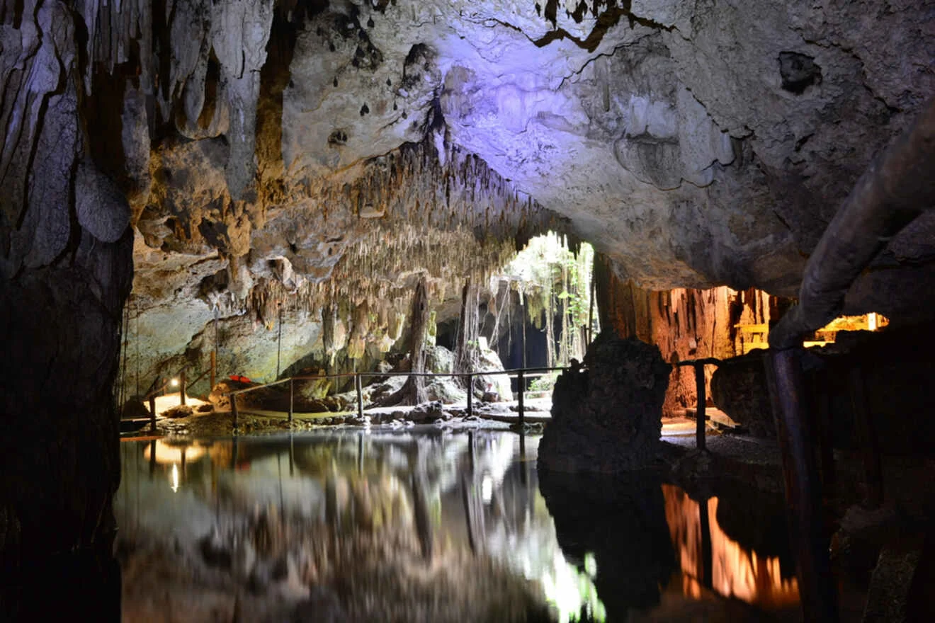 A cave interior with stalactites and stalagmites is illuminated by colored lights, reflecting on a still body of water. A wooden pathway is visible on the left.