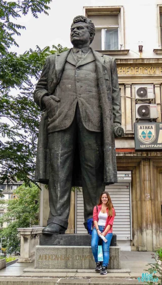 The writer of the post sits on a ledge beside a large statue of a man in a suit and overcoat, with trees and a building in the background.