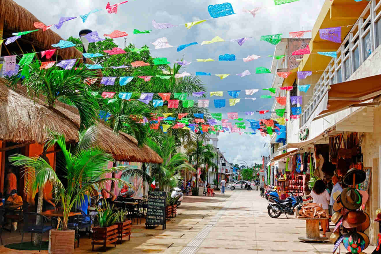 Colorful flags hang above a lively street with shops, palm trees, and outdoor seating under thatched roofs.