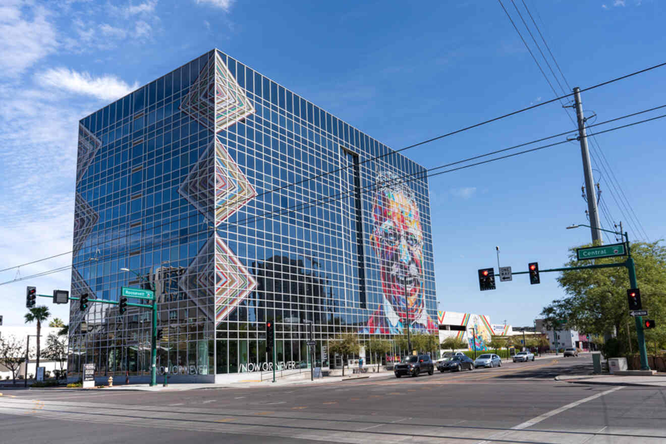 A modern glass building with geometric and colorful mural designs on the facade, located at an intersection with traffic lights and power lines.