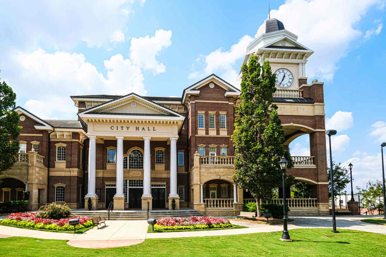 A large brick city hall building with a clock tower and columns, surrounded by trees and flowerbeds under a partly cloudy sky.