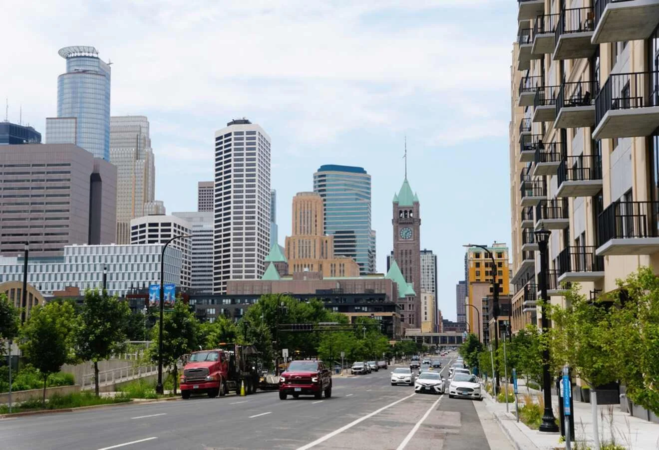 Street view of a cityscape with modern and historic buildings, including a clock tower. Cars are on the road and trees line the street.