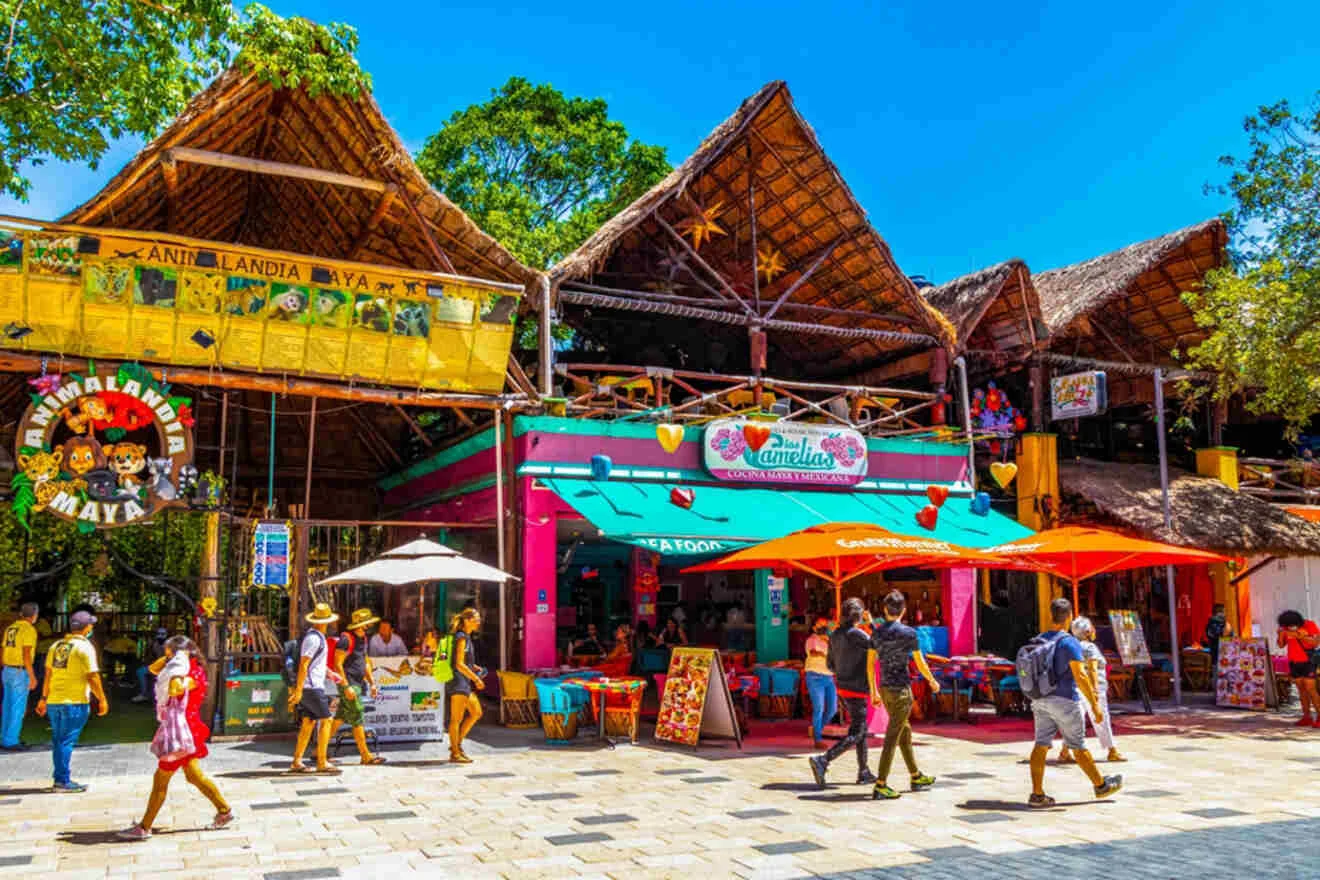 A vibrant outdoor market with thatched roofs, colorful stalls, and people walking along the stone pathway under a sunny sky.