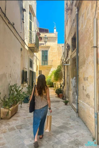 The writer of the post walks down a narrow, sunlit alley with old buildings, carrying a shopping bag and wearing a sleeveless top and denim skirt.