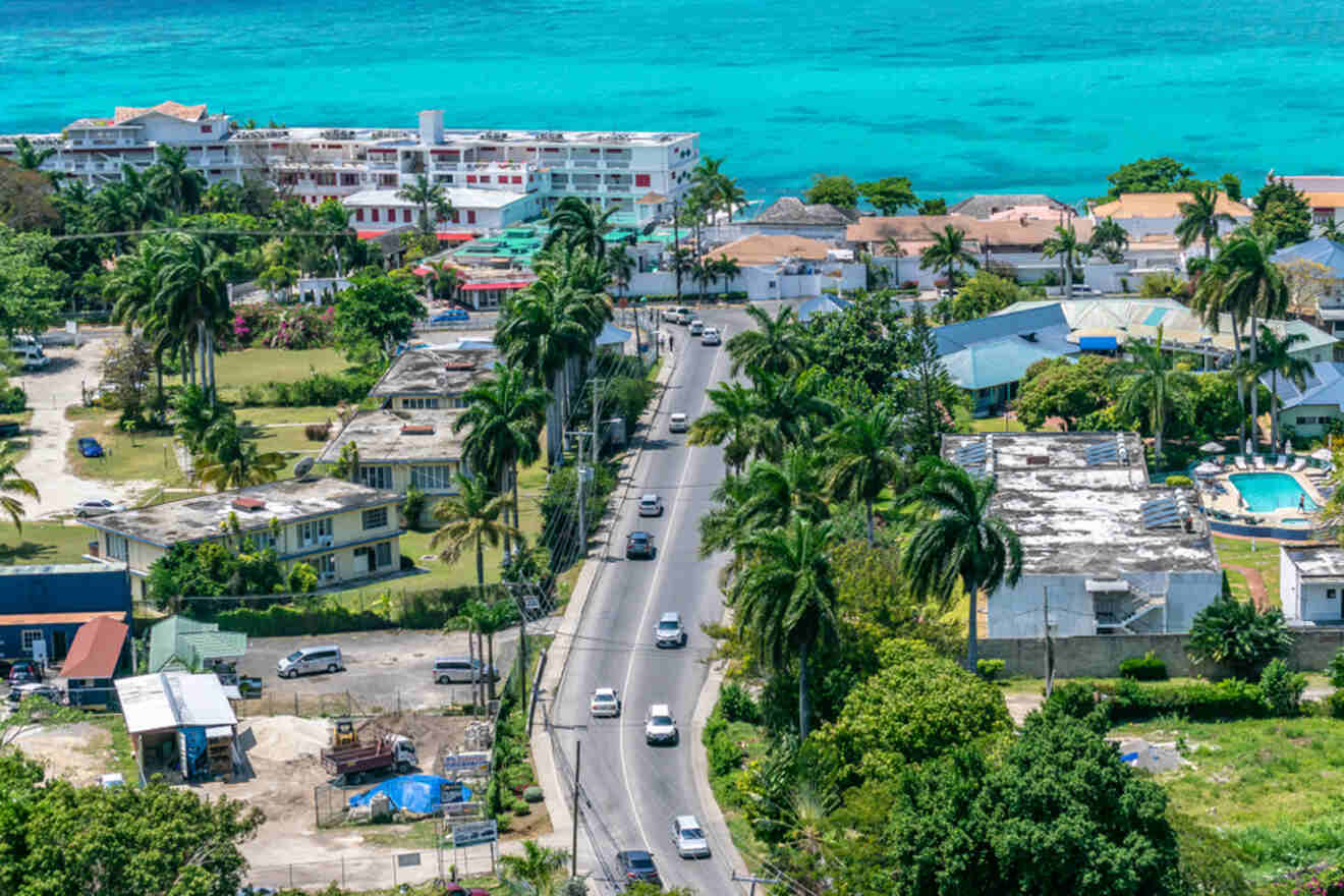 Aerial view of a coastal town with a road lined by palm trees, buildings, and parked cars. The turquoise ocean is visible in the background.