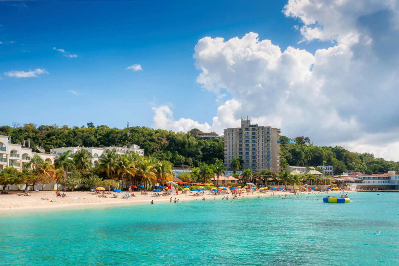 A scenic beach with turquoise water, people sunbathing, colorful umbrellas, and buildings surrounded by greenery under a blue sky with white clouds.