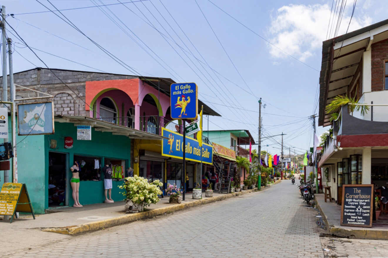 A street scene in a small town with colorful buildings, shops, and overhead power lines. A sign reads "El Gallo mas Gallo" and a few people are visible on the sidewalk.