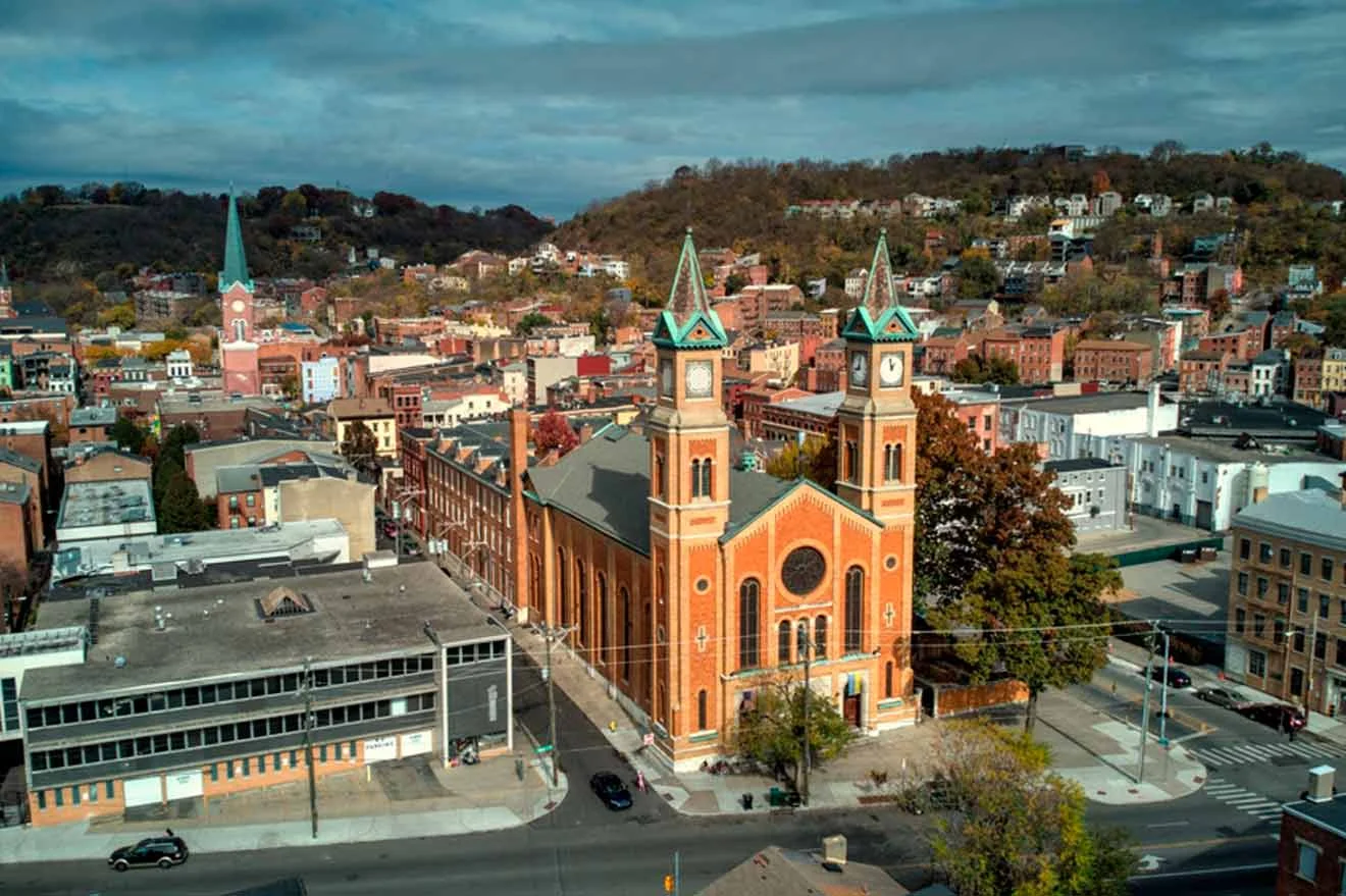 Aerial view of a town with a prominent brick church featuring twin spires. Surrounding buildings and a tree-covered hill are visible under a cloudy sky.