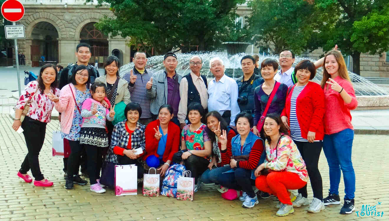 A group of people with The writer of the post poses for a photo in front of a fountain in a paved area with trees and buildings in the background.