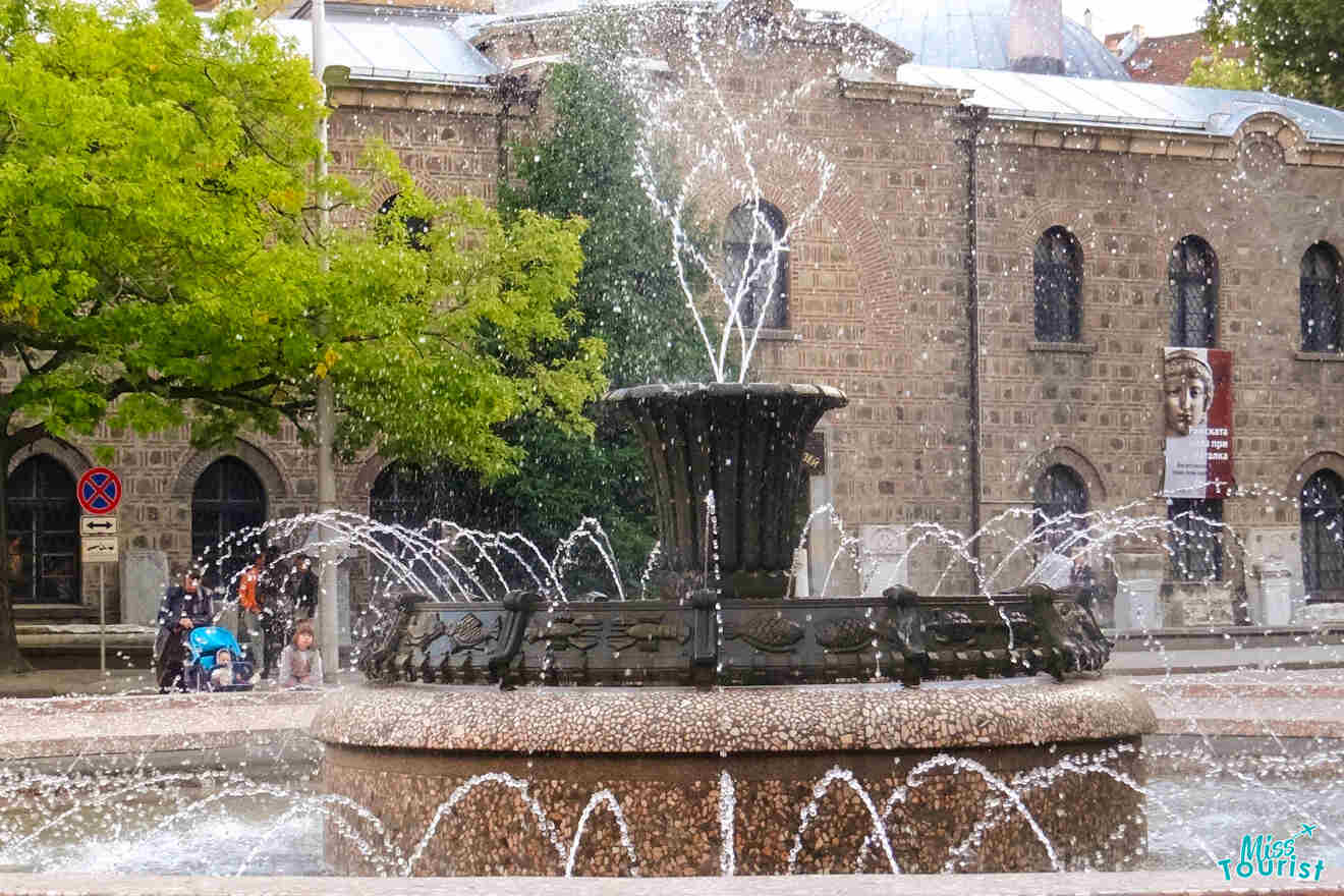 A decorative fountain with water jets is situated in front of a stone building. A tree is on the left, and a sign and stroller are visible nearby.