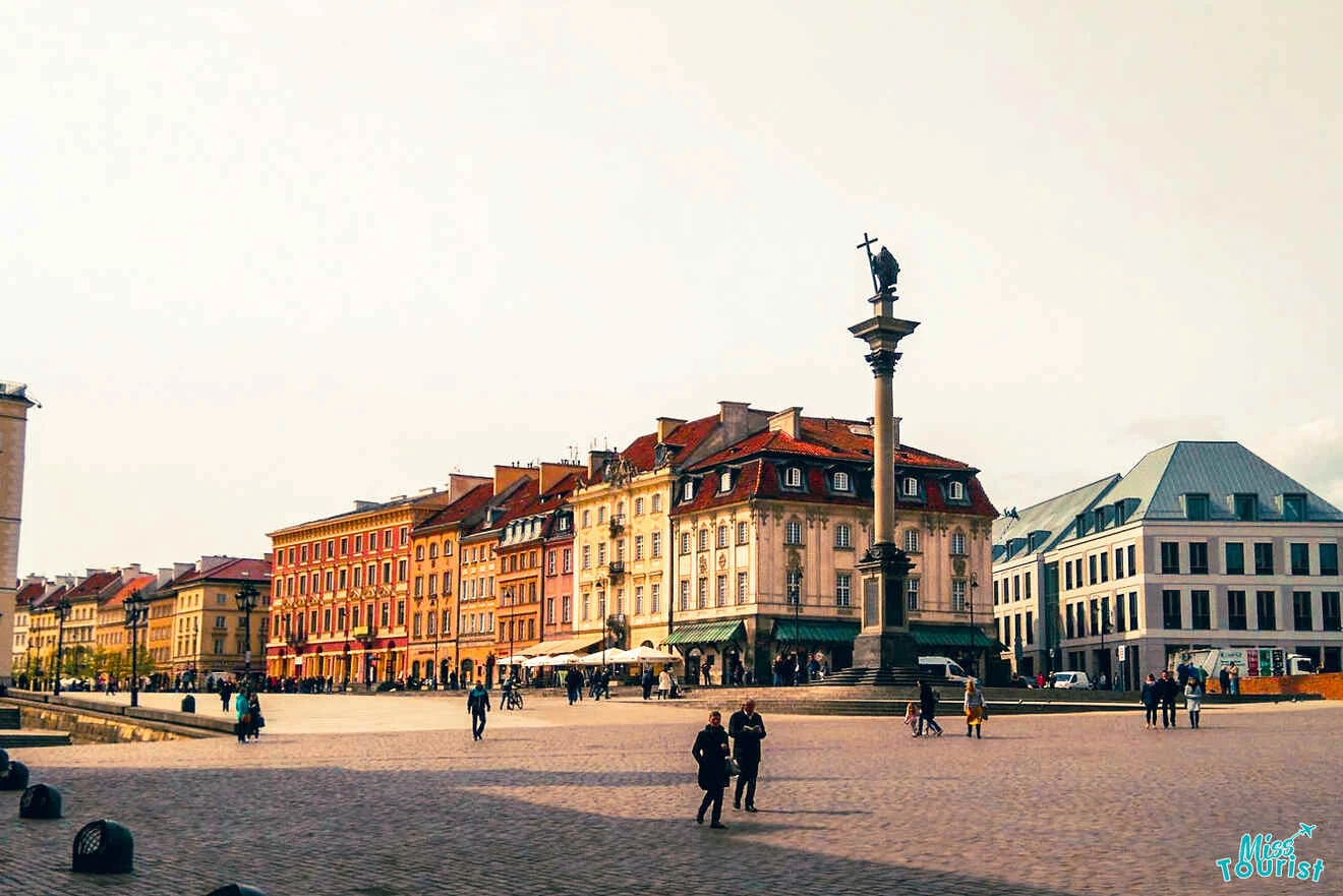 A city square with colorful historic buildings and a tall column topped with a statue. People are walking and gathering in the open space.