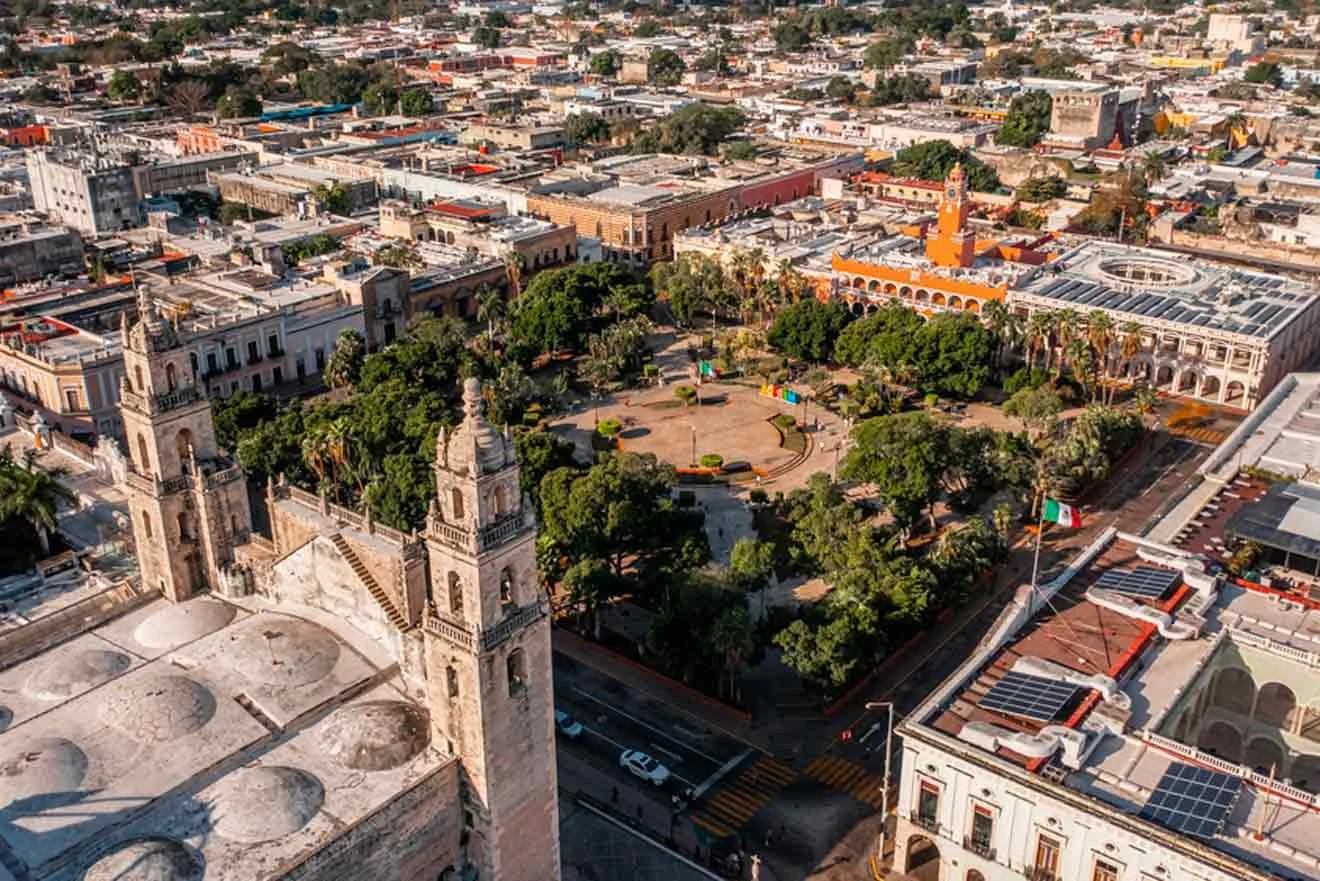 Aerial view of a city square with greenery, surrounded by historic buildings and streets, including a church with towers in the foreground.