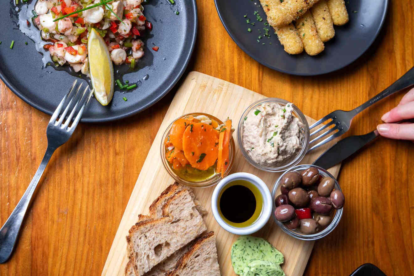 A wooden board with bread, dipping oil, seasoned carrots, a creamy spread, and olives. Plates of shrimp and fried sticks are also visible on the table.