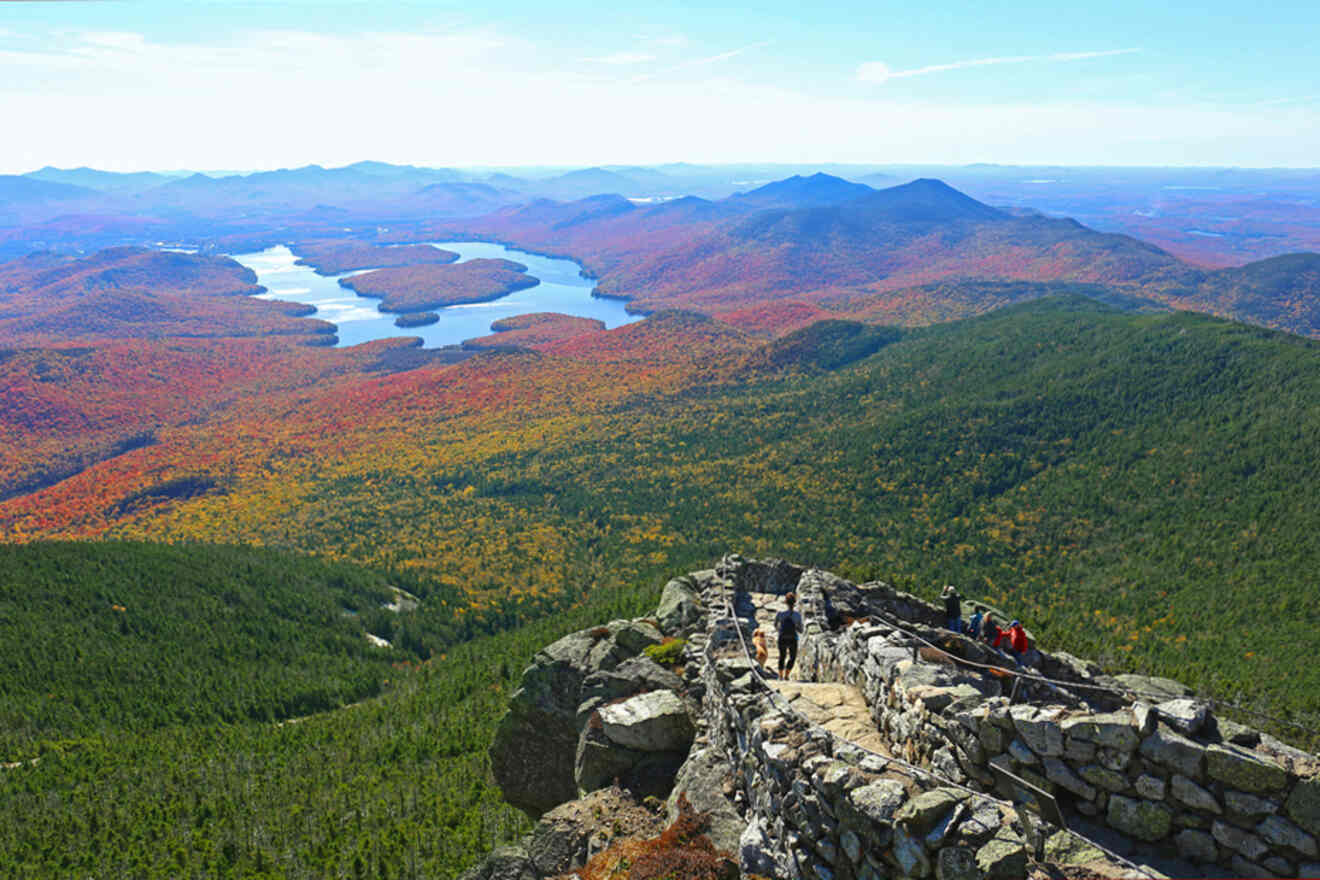 Scenic view from a rocky mountaintop overlooking a winding river and a vast landscape of colorful autumn foliage. Several people are visible on the stone pathway in the foreground.