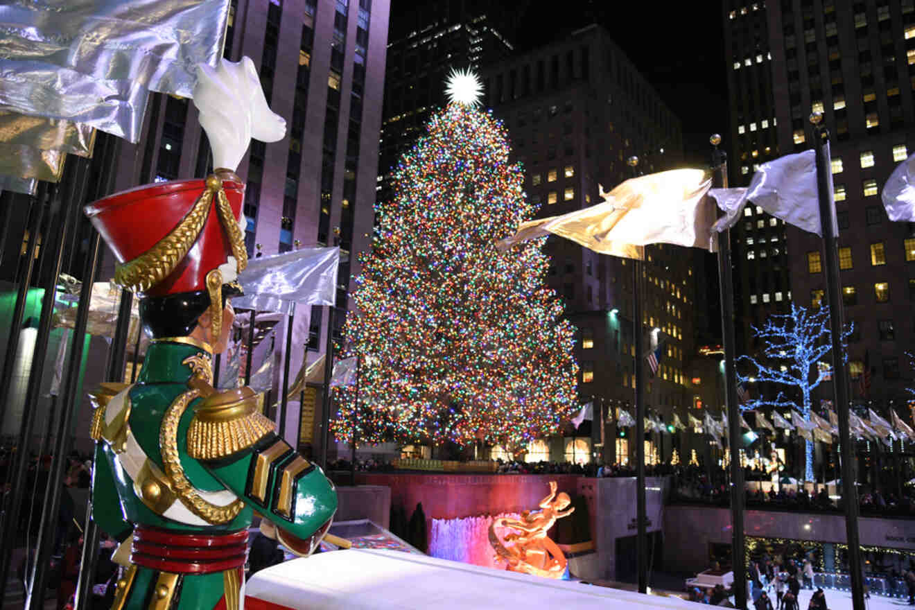 A large Christmas tree decorated with colorful lights stands in a plaza at night, with a toy soldier statue in the foreground and flags waving nearby.