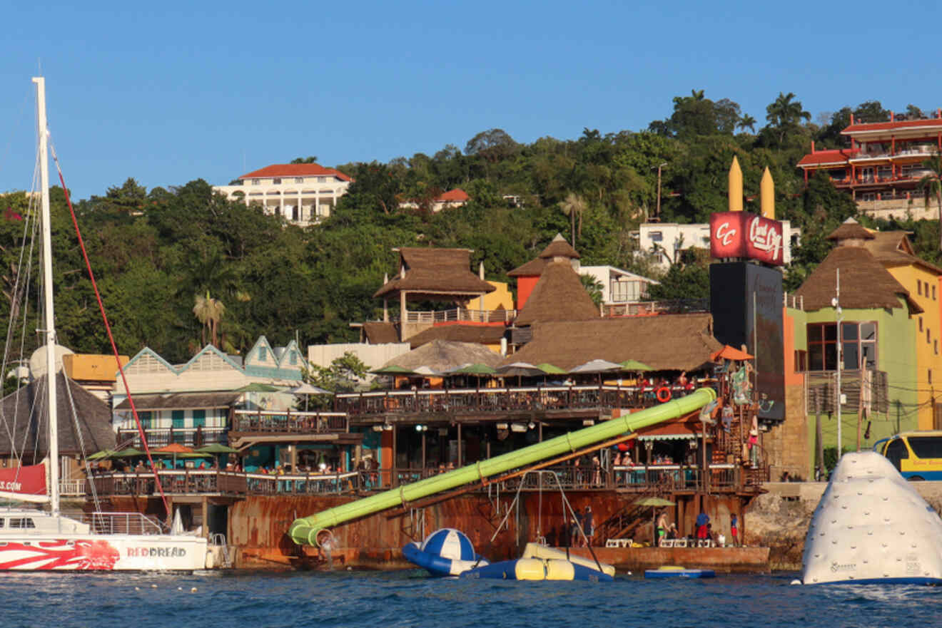 A waterfront view of a lively restaurant with a large waterslide, inflatable structures, and sailboats docked nearby, surrounded by lush greenery and colorful buildings.