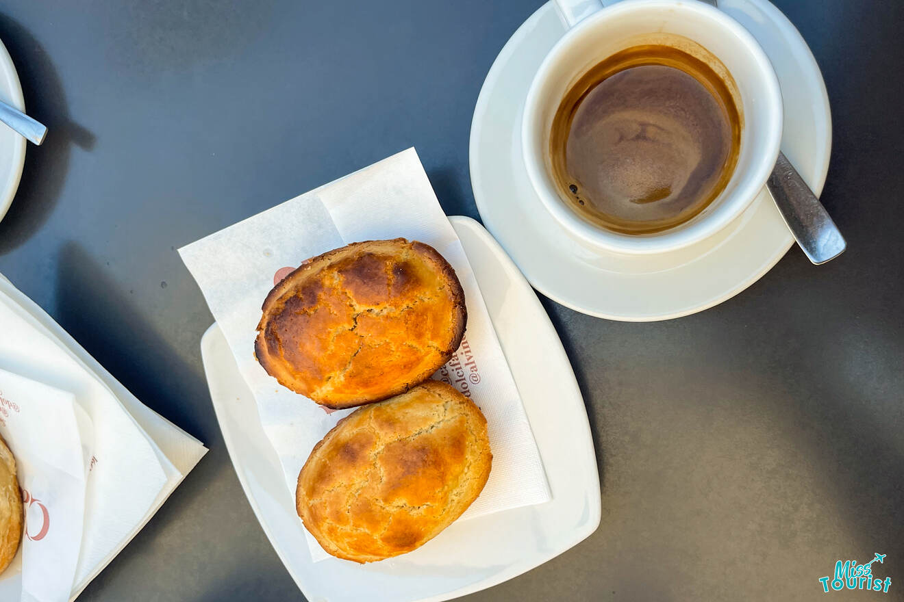 Two golden-brown pastries on a plate with a paper napkin next to a cup of espresso on a dark tabletop.