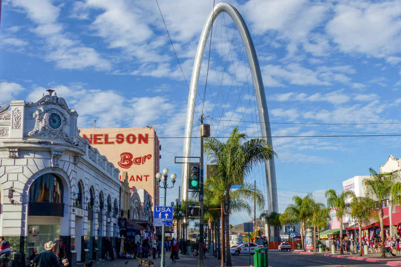 Street scene with a large arch, palm trees, and colorful buildings under a blue sky in a bustling urban area.