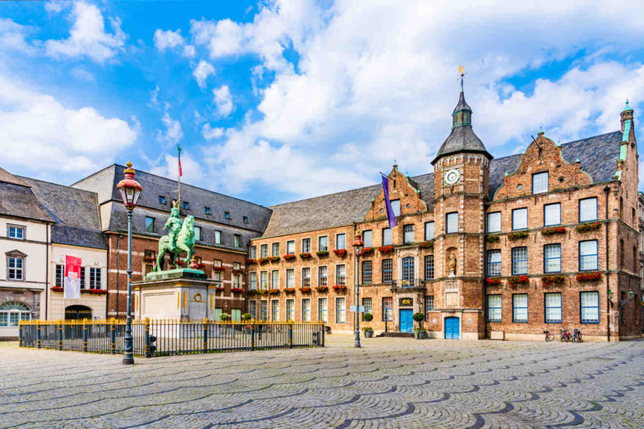 Historic building with a clock tower and a statue of a rider on horseback in a cobblestone square. Building adorned with flags and flowers. Blue sky with some clouds overhead.