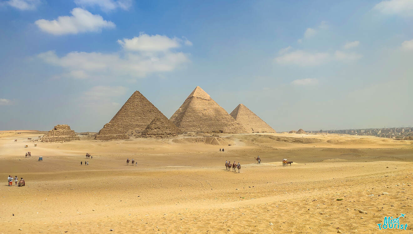 Three pyramids stand on a sandy desert under a blue sky with scattered clouds. Tourists and camels are visible in the foreground.