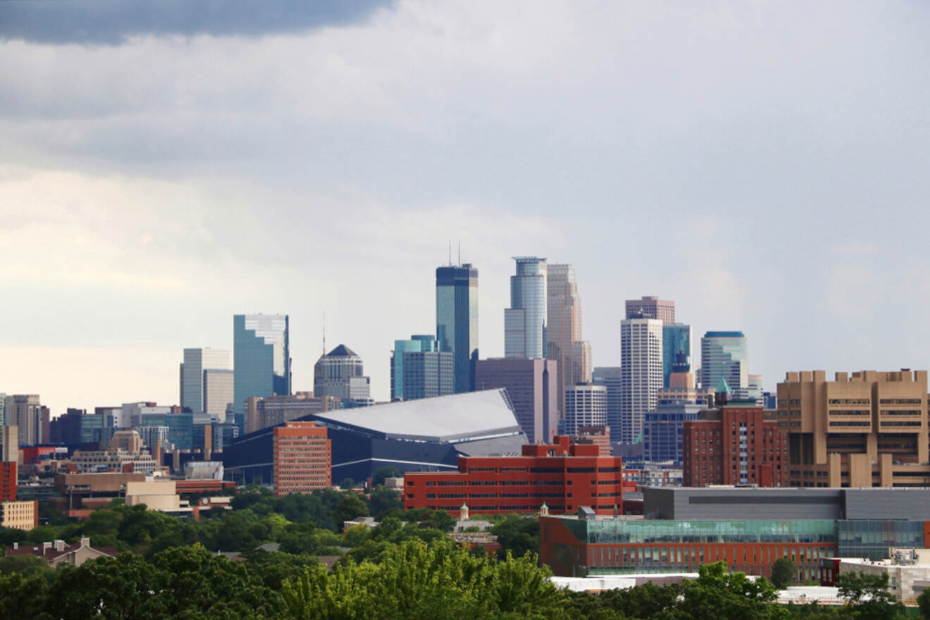 Skyline of a city with modern skyscrapers and a prominent stadium, under a cloudy sky. Greenery and urban buildings are in the foreground.