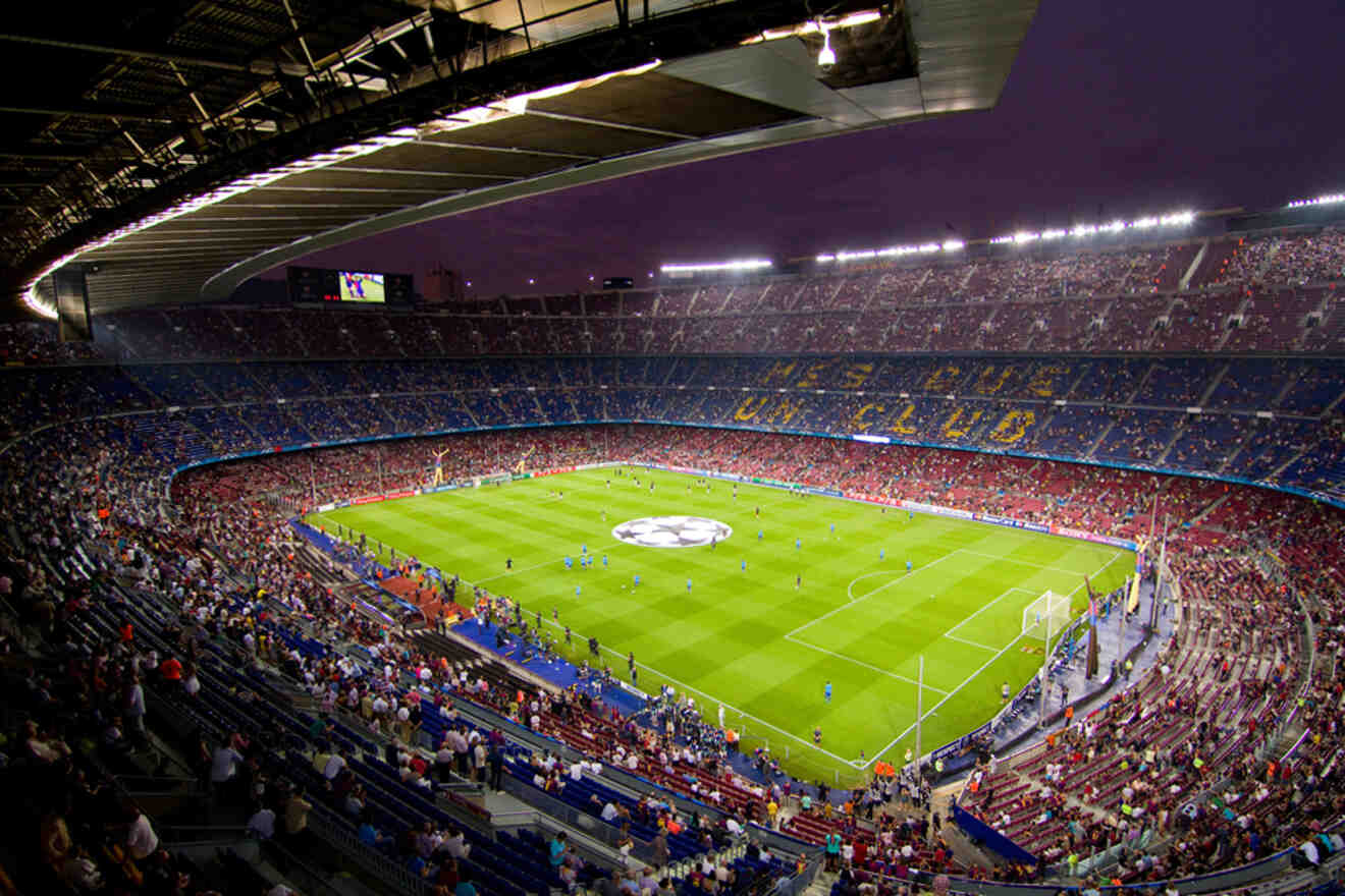 A packed football stadium with bright green pitch, players warming up, and a large crowd in the stands during an evening match.