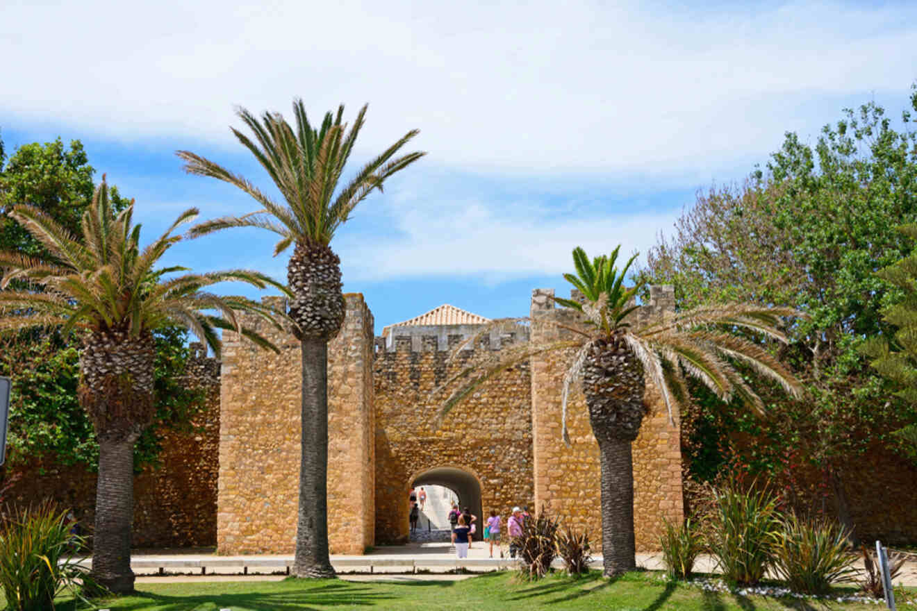 The stone castle wall in Lagos, Portugal, boasts an arched entrance flanked by palm trees and lush greenery. Under the blue sky, people stroll near the entrance.