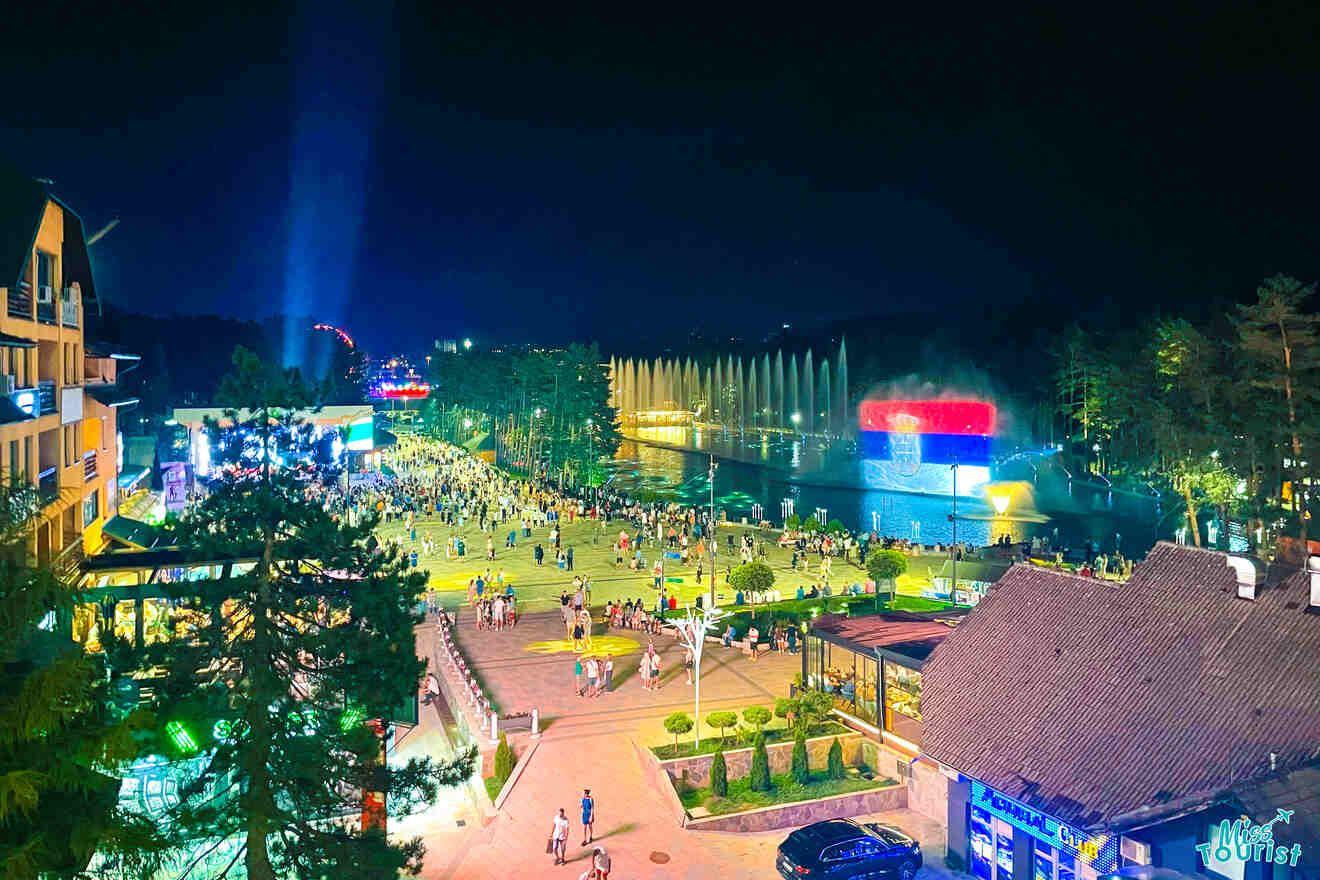 A vibrant night scene unfolds as a crowd gathers near an illuminated water fountain show in an urban park, a highlight of any Serbia itinerary. Buildings and trees beautifully frame the spectacle, creating an enchanting atmosphere.