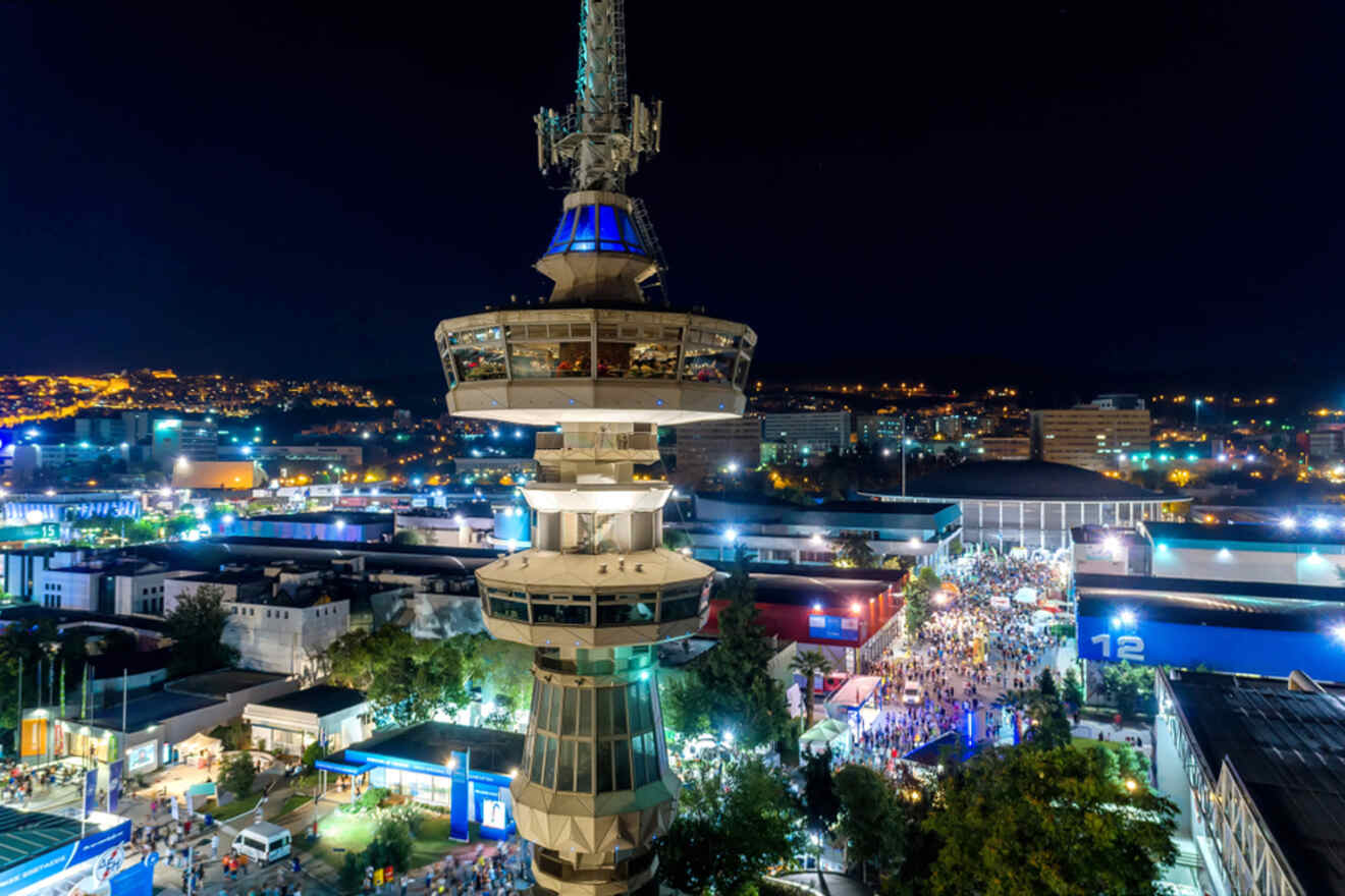 A tall tower stands illuminated against a bustling night scene in an urban area, with bright city lights and crowds visible below.