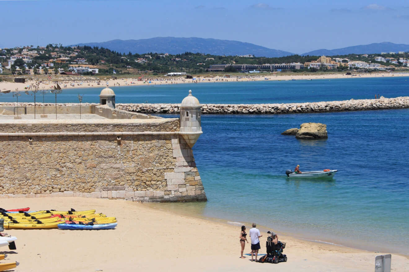 Beach scene with a stone fort on the left, kayaks on sand, and a family with a stroller walking nearby. A person in a small boat is on the water, with Lagos, Portugal's townscape and mountains as a picturesque backdrop.