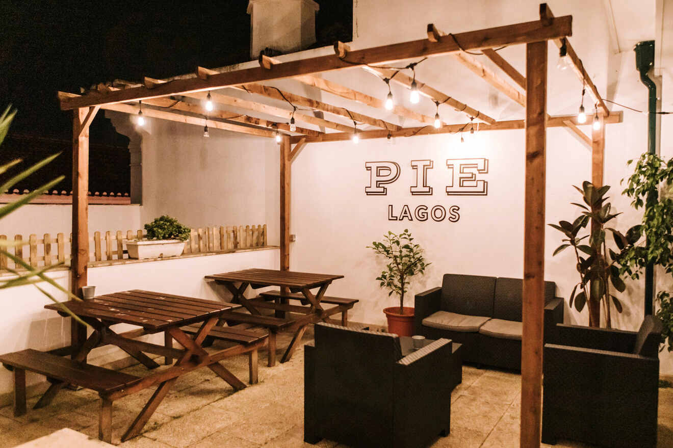 Outdoor patio with wooden benches, string lights, and plants, featuring a sign that reads "PIE LAGOS.