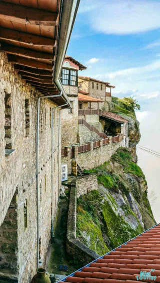 A stone building with red-tiled roof sits on a cliff edge, overlooking a cloudy sky. Wooden railings line the balconies.