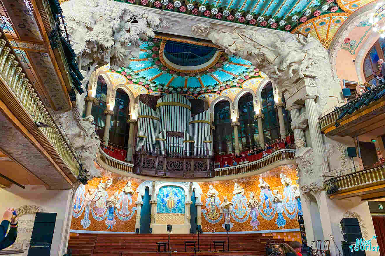 Interior view of a concert hall with ornate architecture, featuring a colorful stained glass ceiling, intricate sculptures, and a large organ.