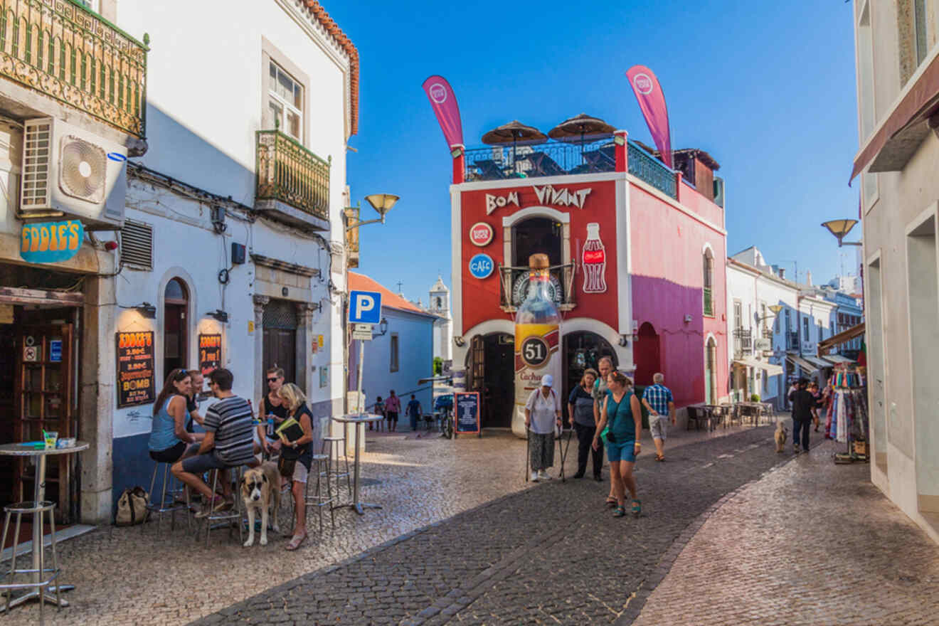 People walk and sit outside cafés on a cobblestone street lined with colorful buildings under a clear blue sky.